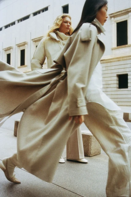 Two women in long coats walk on a city sidewalk with neutral stone buildings. One woman is mid-stride, coat flowing open.