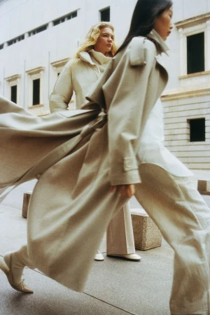 Two women in long coats walk on a city sidewalk with neutral stone buildings. One woman is mid-stride, coat flowing open.