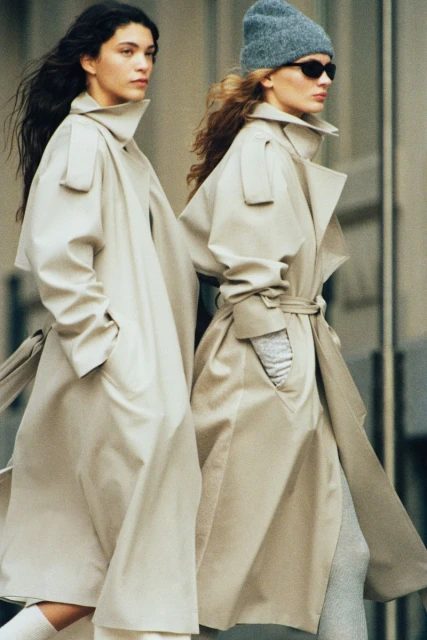 Two women in trench coats walk outdoors, one wears a beanie and sunglasses, showcasing a blend of classic and modern fashion.