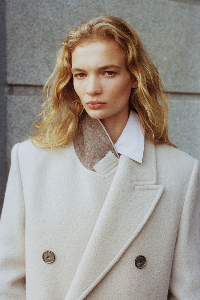 Close-up of a blonde woman in a cream wool coat and white shirt, looking at the camera against a textured gray wall backdrop.