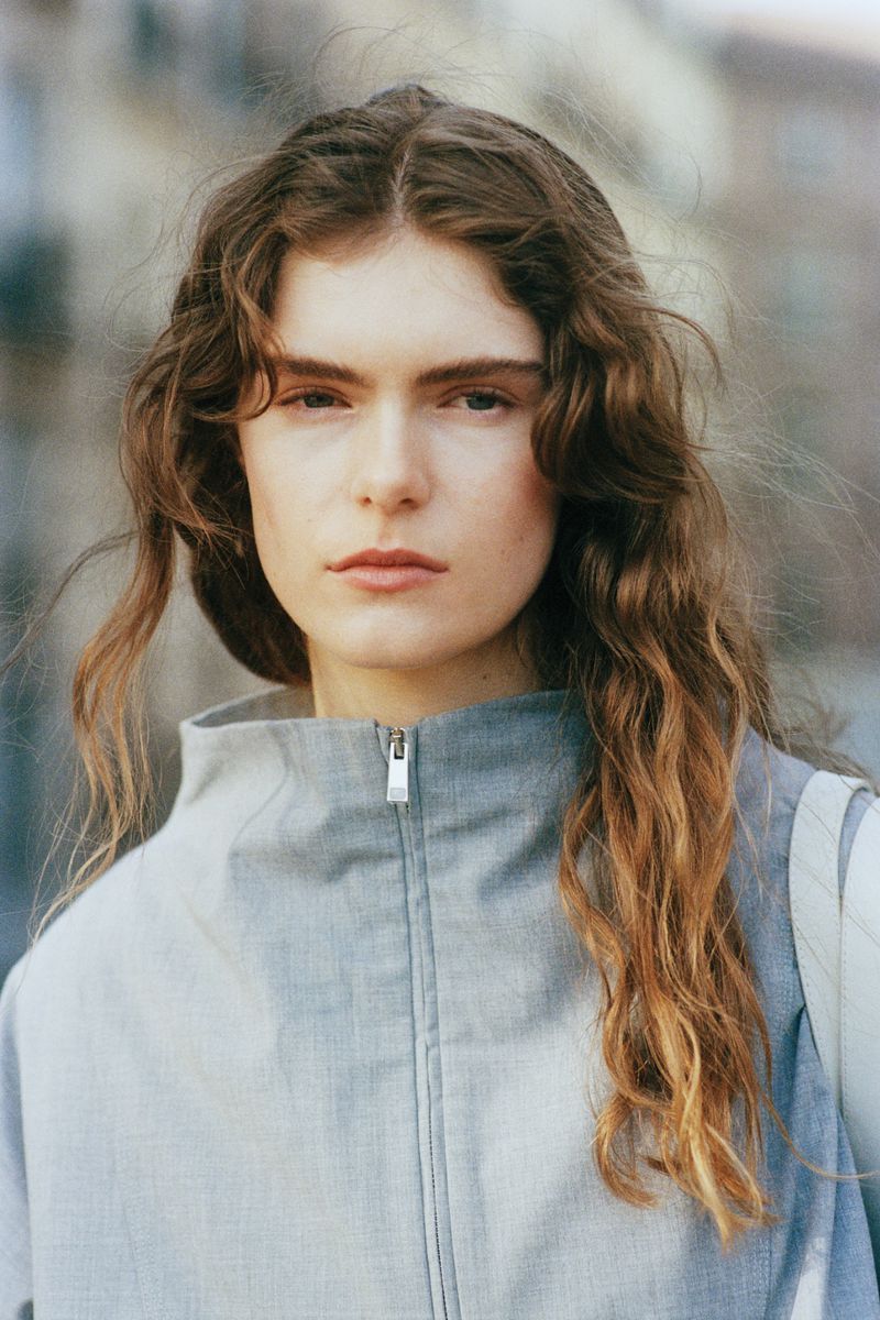 Portrait of a female model with long wavy brown hair wearing a grey zip collar top outdoors