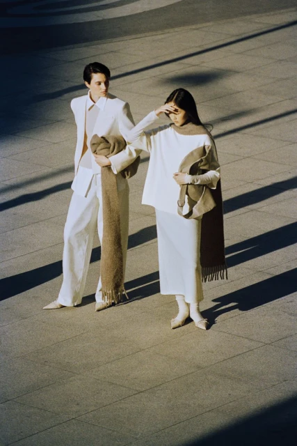 Two fashion models in neutral-tone outfits pose on a concrete surface with linear shadow patterns in bright daylight, each holding a scarf.