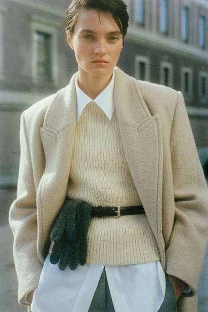 A stylish woman in a beige coat, cream sweater, and green trousers stands outdoors in front of a stone building on a sunny day.