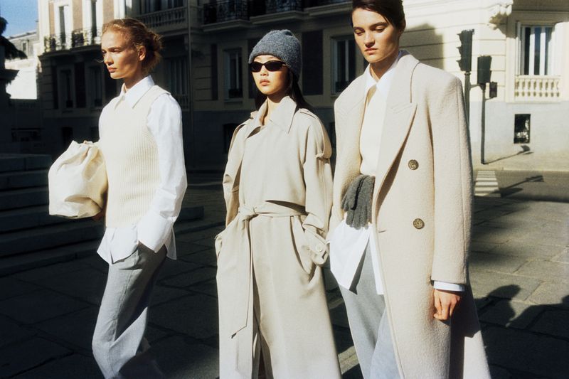 Three adult models in stylish beige overcoats walk on an urban street with buildings in the background, basking in bright daylight.