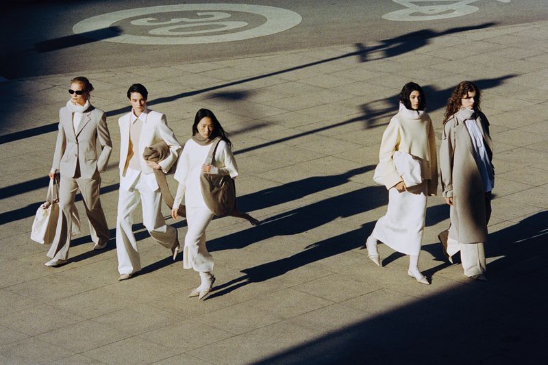 Five models walking across a paved public square in neutral outfits, cast with long shadows under bright daylight.