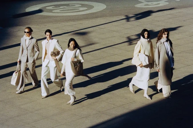 Five models walking across a paved public square in neutral outfits, cast with long shadows under bright daylight.