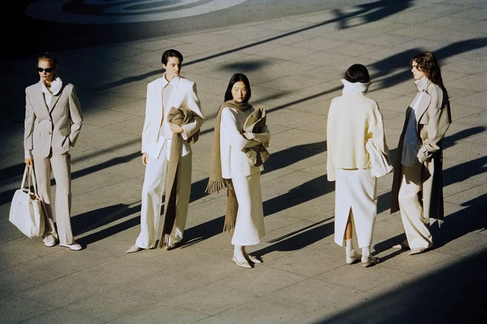 Five models in neutral-toned apparel standing on a paved outdoor square casting long diagonal shadows
