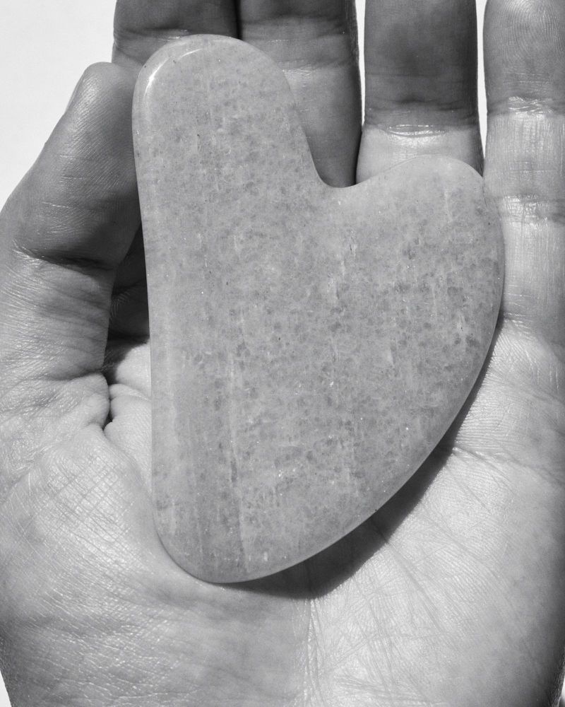 Close-up of a hand holding a gua sha facial massage tool made from light-colored stone, set against a bright, neutral background.