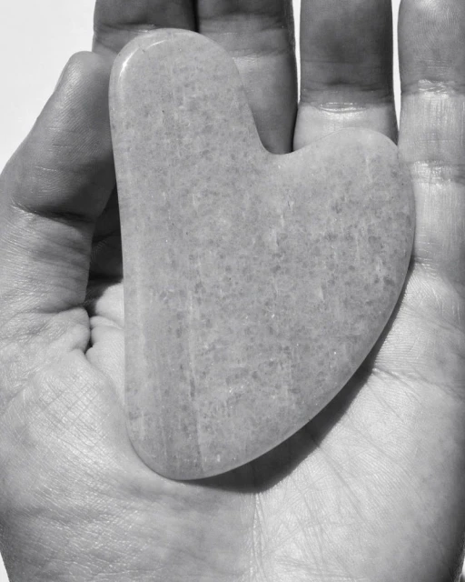 Close-up of a hand holding a gua sha facial massage tool made from light-colored stone, set against a bright, neutral background.
