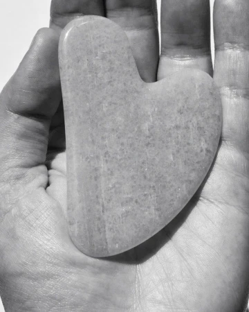 Close-up of a hand holding a gua sha facial massage tool made from light-colored stone, set against a bright, neutral background.