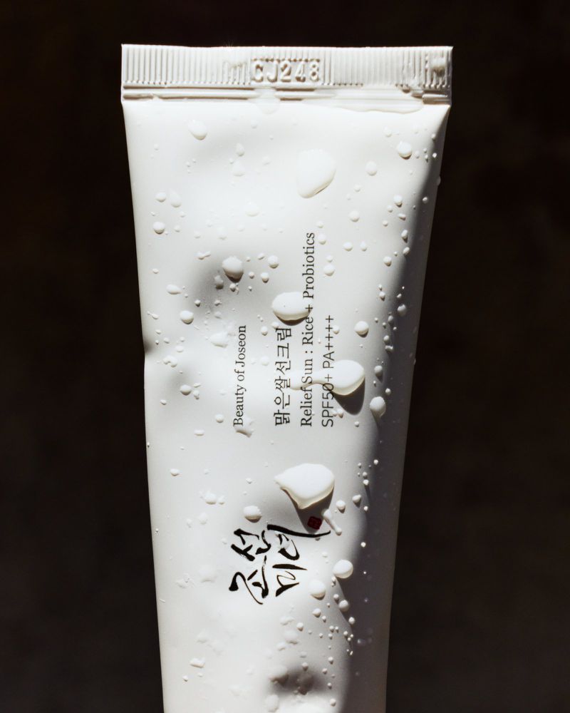 Close-up of a white tube of Beauty of Joseon sunscreen with water droplets against a dark background, product code CJ248 visible.