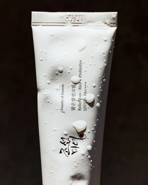 Close-up of a white tube of Beauty of Joseon sunscreen with water droplets against a dark background, product code CJ248 visible.