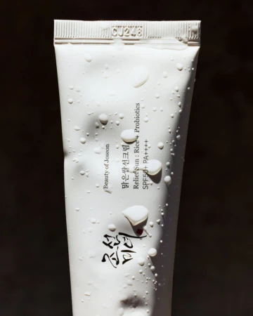 Close-up of a white tube of Beauty of Joseon sunscreen with water droplets against a dark background, product code CJ248 visible.