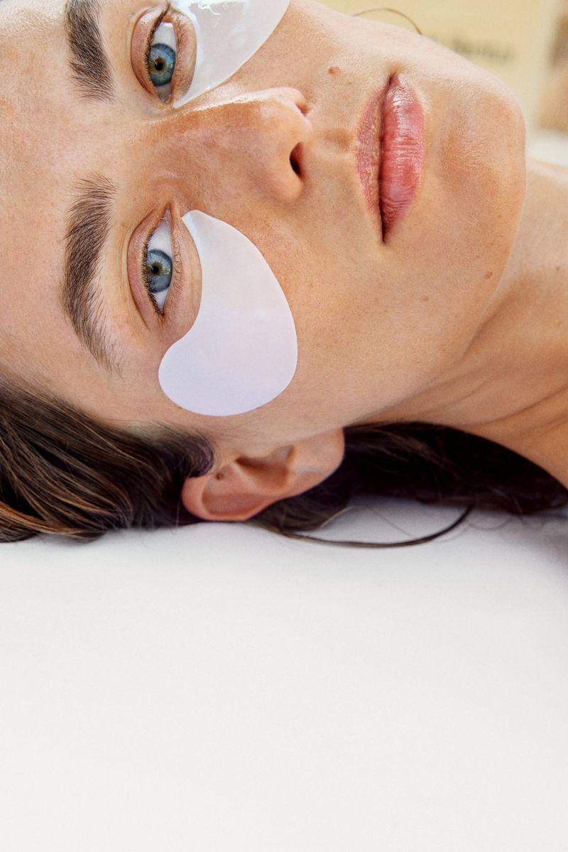 Close-up of a light-skinned woman with blue eyes, lying down with eye patches, looking at the camera with a serene expression.
