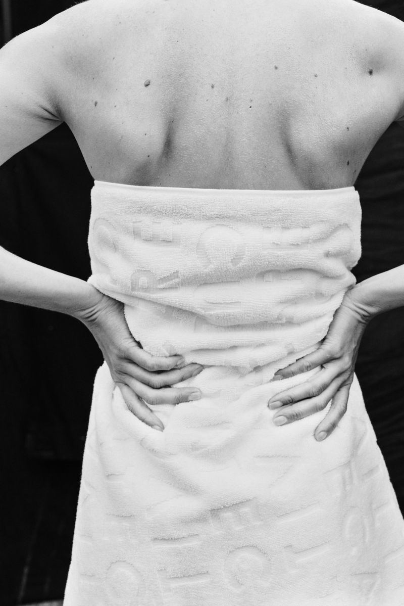 Black and white shot of an adult female from the back, wrapped in a textured white towel with hands on hips, against a dark background.