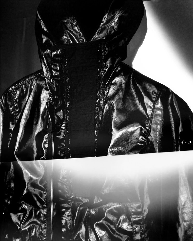 Close-up of a black, hooded, shiny jacket with zipper, under studio lighting, creating a dramatic and high-contrast fashion product shot.