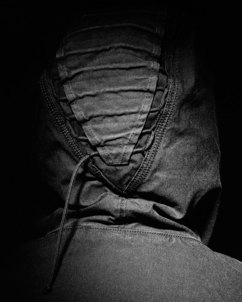 Close-up of a person wearing a dark hooded jacket, seen from the back with the hood up, set against a black backdrop.