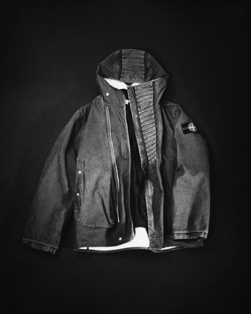 Studio shot of a gray Stone Island hooded jacket, slightly open, against a dark backdrop with a visible logo patch on the left sleeve.