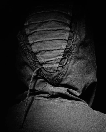 Rear view of a person wearing a dark hooded jacket with paneling and drawstrings against a solid black background.