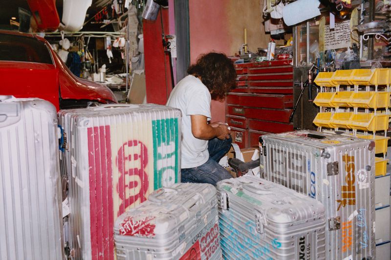 Adult male kneeling amidst luggage in cluttered garage, examining items in daylight. Red car visible in background.