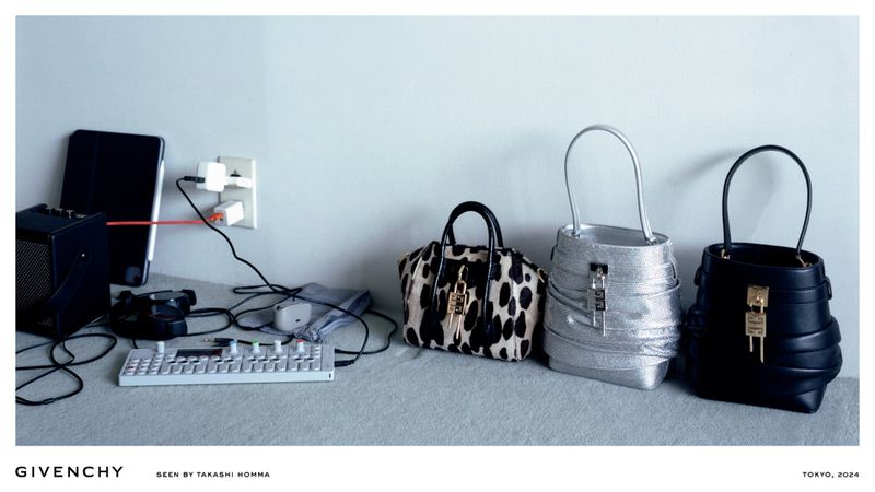 Fashion editorial shot of three Givenchy handbags arranged next to electronic devices and charging cables on a gray surface.