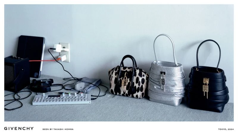 Fashion editorial shot of three Givenchy handbags arranged next to electronic devices and charging cables on a gray surface.