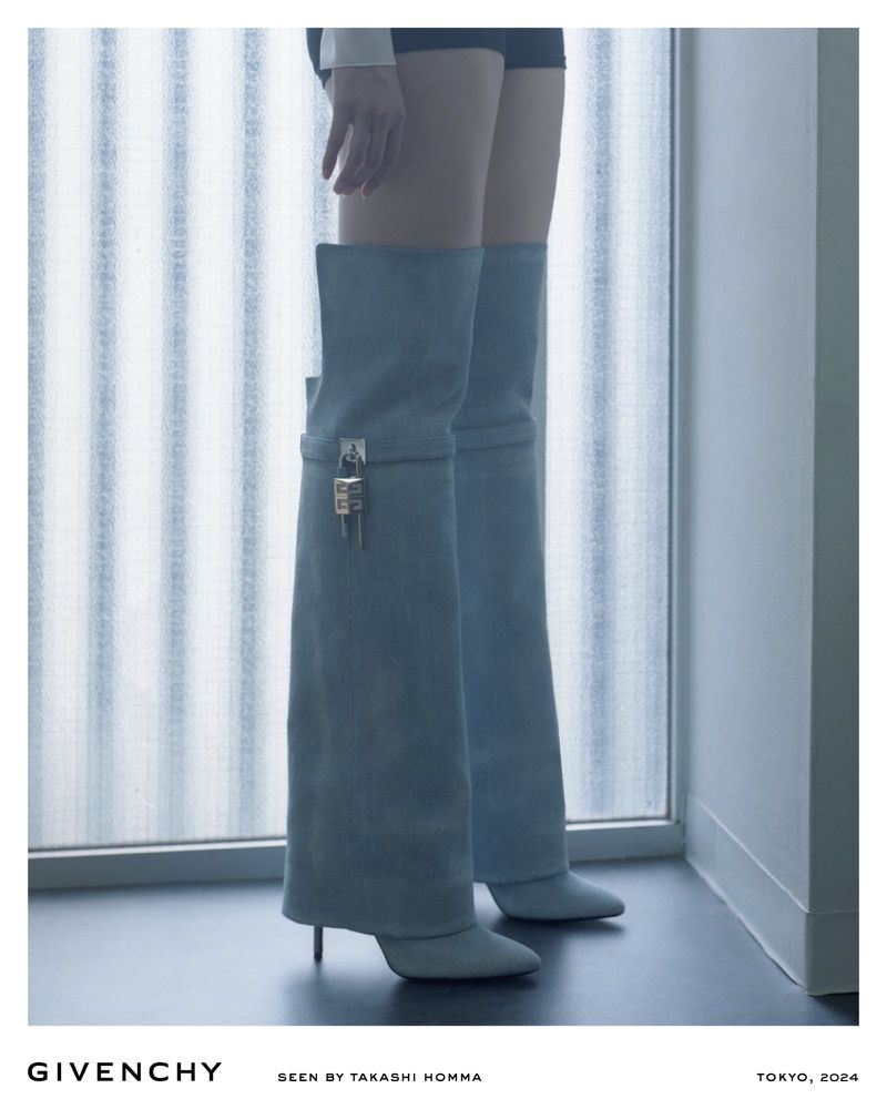 Fashion editorial shot of an adult woman wearing light blue thigh-high lock boots standing in front of a window.