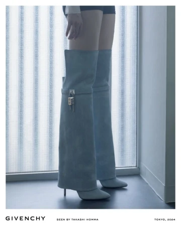 Fashion editorial shot of an adult woman wearing light blue thigh-high lock boots standing in front of a window.