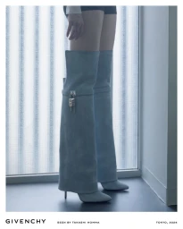 Model wearing tall blue denim Shark Lock boots with silver padlock detail standing in front of a window