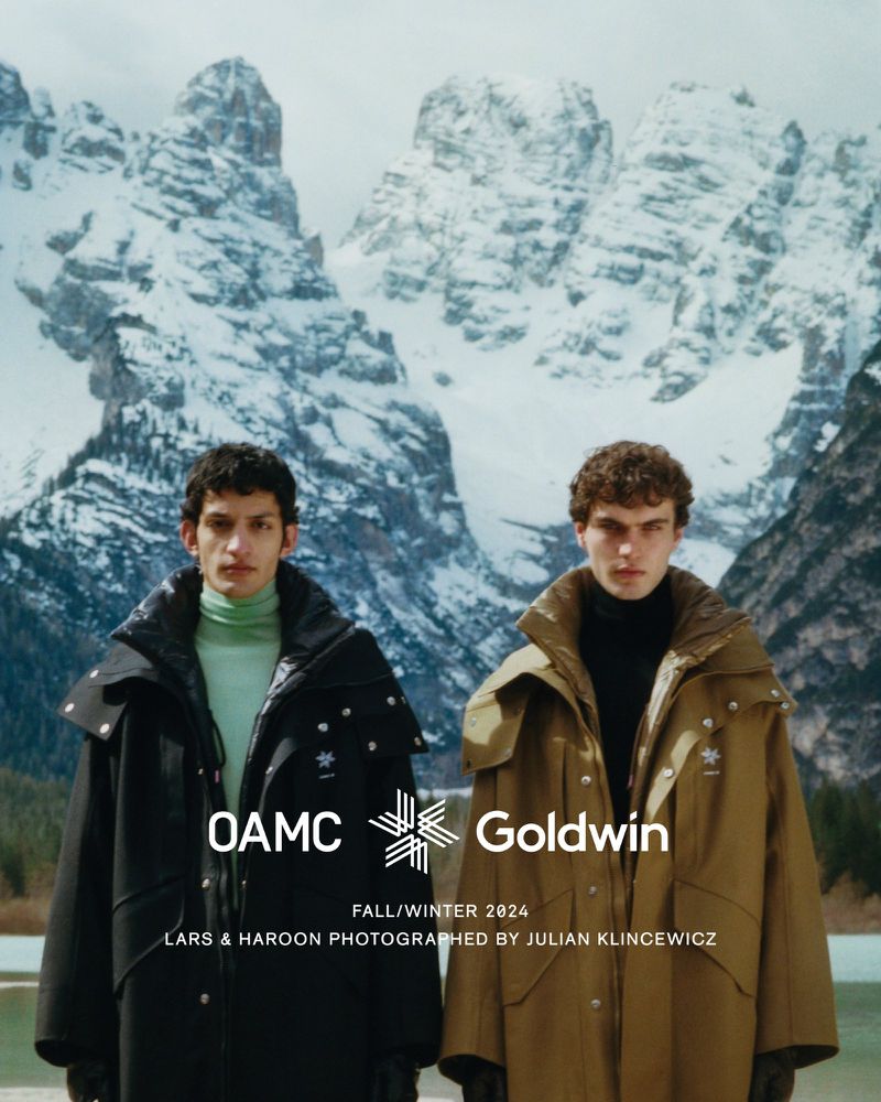 Two men in long winter coats pose in front of snow-covered mountains. The image promotes a collaboration between OAMC and Goldwin.