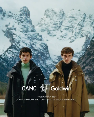 Two men in long winter coats pose in front of snow-covered mountains. The image promotes a collaboration between OAMC and Goldwin.