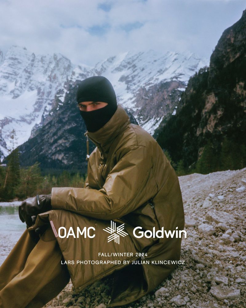Person in black balaclava and khaki coat sits by a stream with snowy mountains in the background in an outdoor fashion shot.