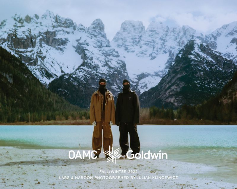Two people in OAMC and Goldwin fall/winter 2024 collection stand in front of a lake with snow-capped mountains in the background.