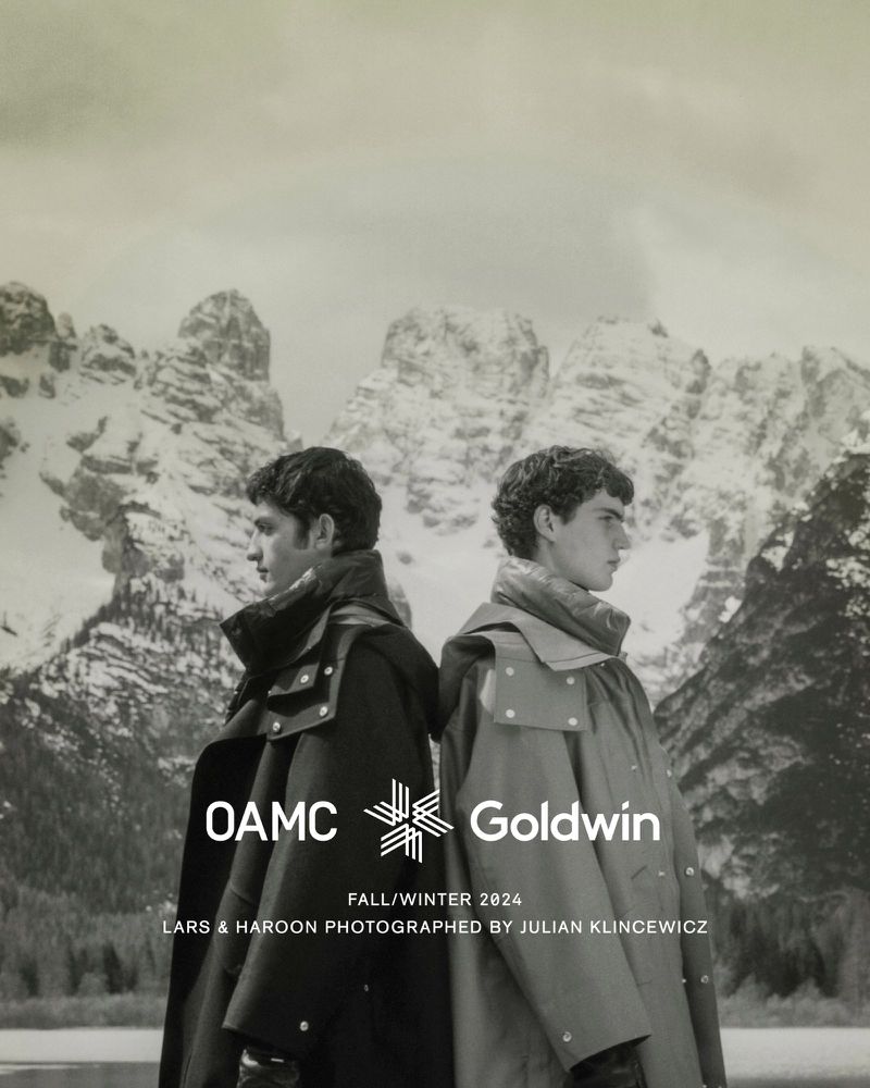 Two men wearing coats stand back-to-back in a promotional shot featuring a snowy mountain range and the OAMC and Goldwin logos.