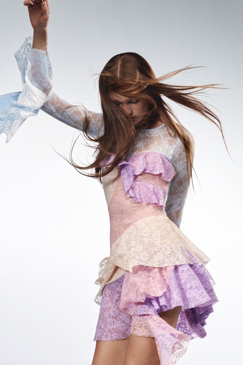 A woman in a pastel-colored lace dress with ruffled details strikes a pose in a fashion studio, showcasing her attire against a neutral backdrop.