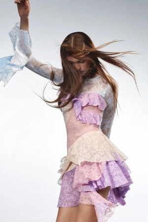 A woman in a pastel-colored lace dress with ruffled details strikes a pose in a fashion studio, showcasing her attire against a neutral backdrop.