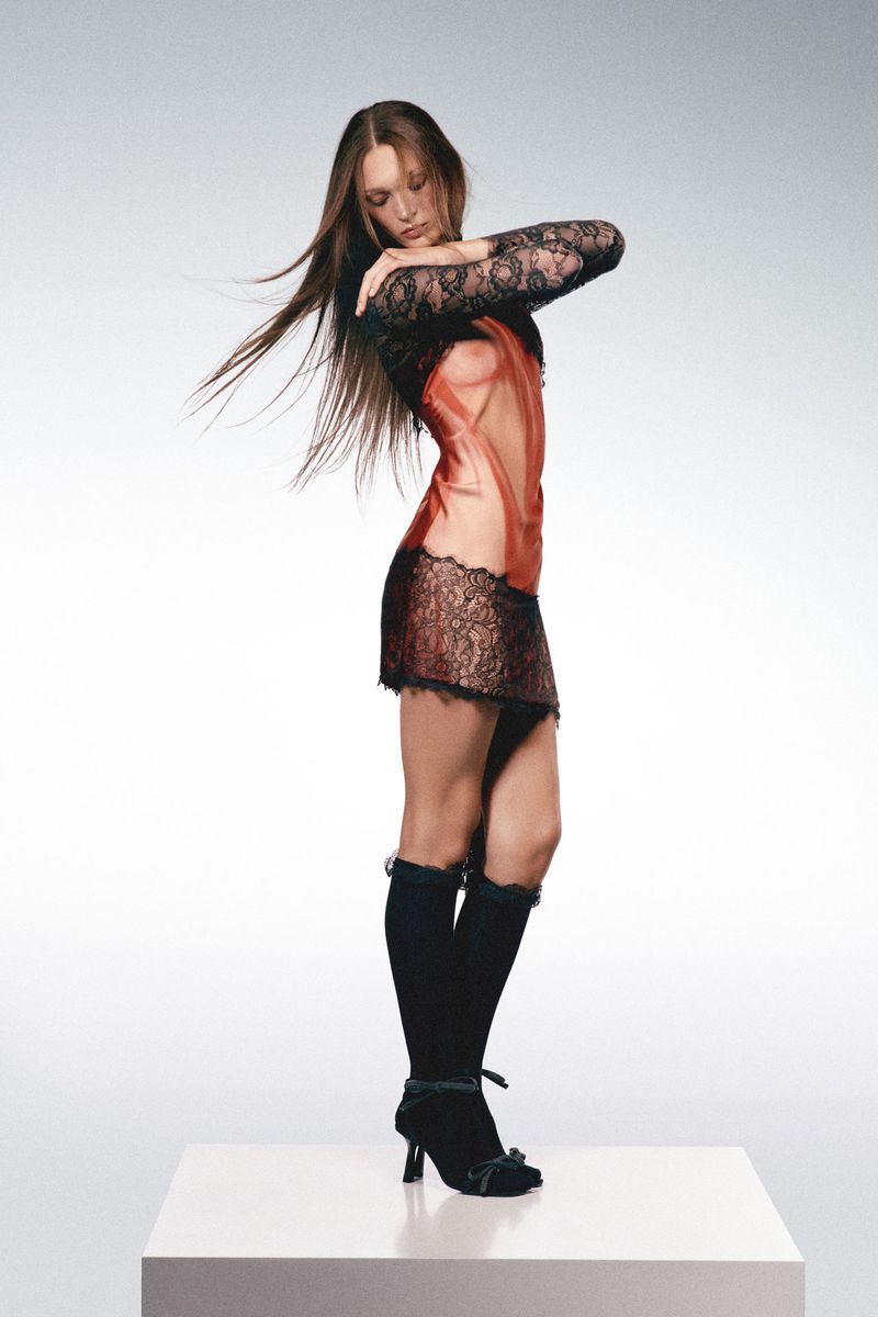 A slender woman in a sheer coral and black lace dress poses on a white platform in a photo studio with a soft gradient background.