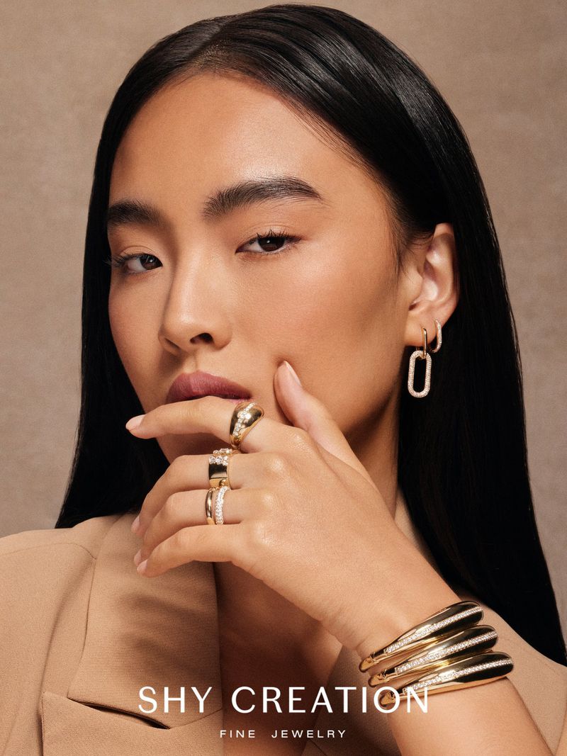 Elegant Asian woman showcases gold and diamond jewelry: rings, bracelet, and earrings against a neutral backdrop in a beauty shot.