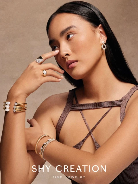 Close-up shot of an Asian woman modeling gold and diamond bracelets and rings, wearing a knit top, against a beige background.