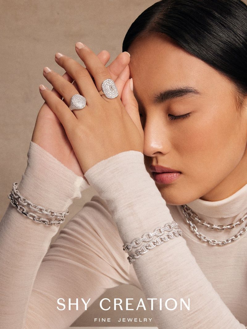 Close-up portrait of a woman wearing diamond rings and chain bracelets against a neutral textured background