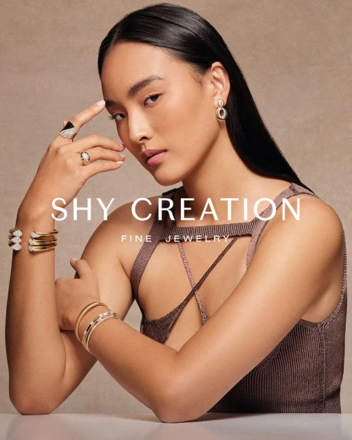 Elegant woman poses wearing gold jewelry, including rings, earrings, and bracelets, against a neutral backdrop in a studio shot.