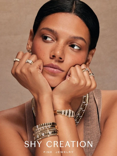 Close-up shot of an adult female model with gold rings, bracelets, and necklace; the model is posing in a studio with a tan backdrop.