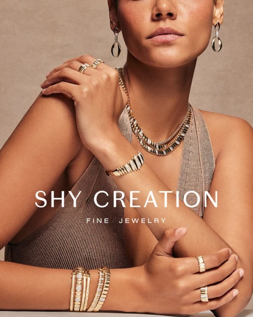 Close-up of a woman showcasing gold jewelry, including a necklace, earrings, rings, and bracelets against a neutral background.