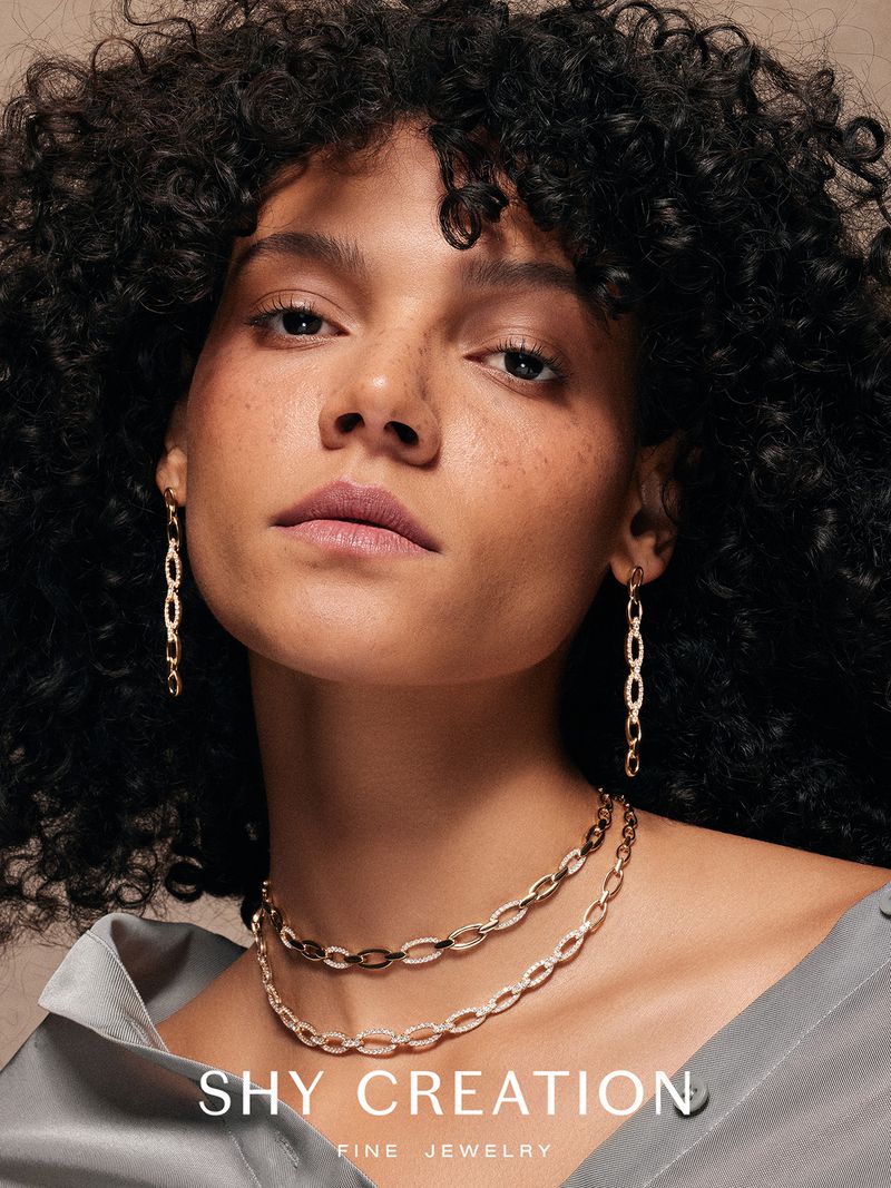 Close-up shot of a brunette woman with gold earrings and layered gold necklaces against a neutral backdrop, showcasing jewelry.