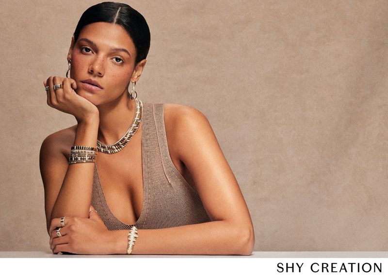 Woman in a tank top poses with an array of jewelry. She has on a necklace, bracelets, rings, and earrings in a studio setting.