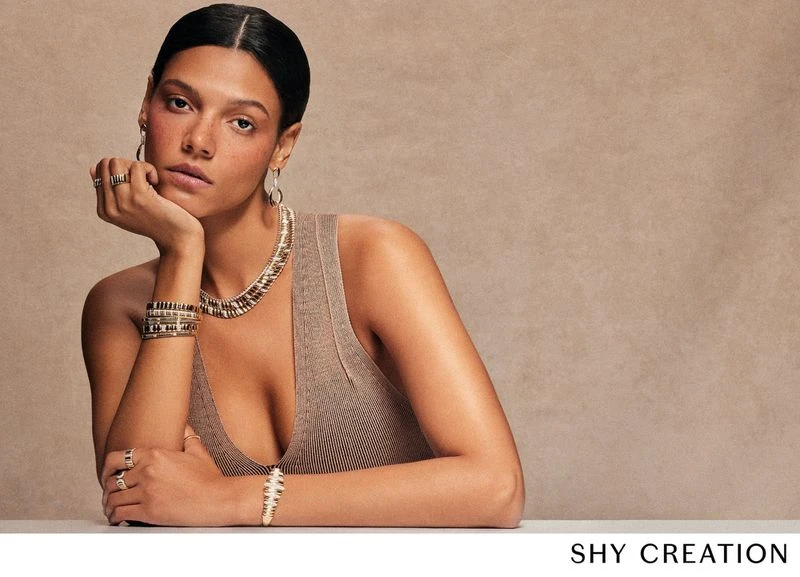 Woman in a tank top poses with an array of jewelry. She has on a necklace, bracelets, rings, and earrings in a studio setting.