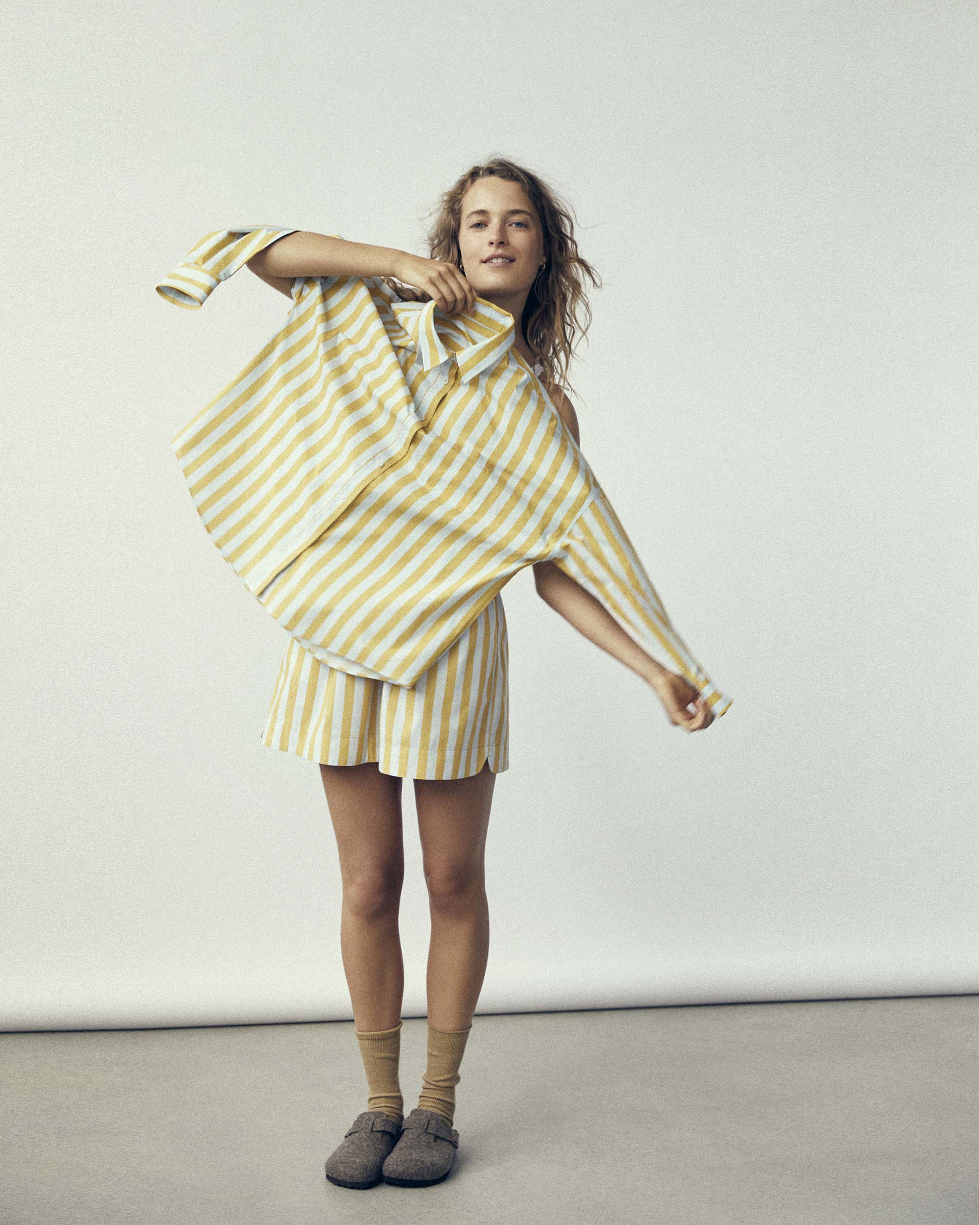 A smiling woman in a striped matching set poses in front of a white background, playfully holding the shirt from the set.