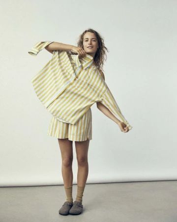 A smiling woman in a striped matching set poses in front of a white background, playfully holding the shirt from the set.
