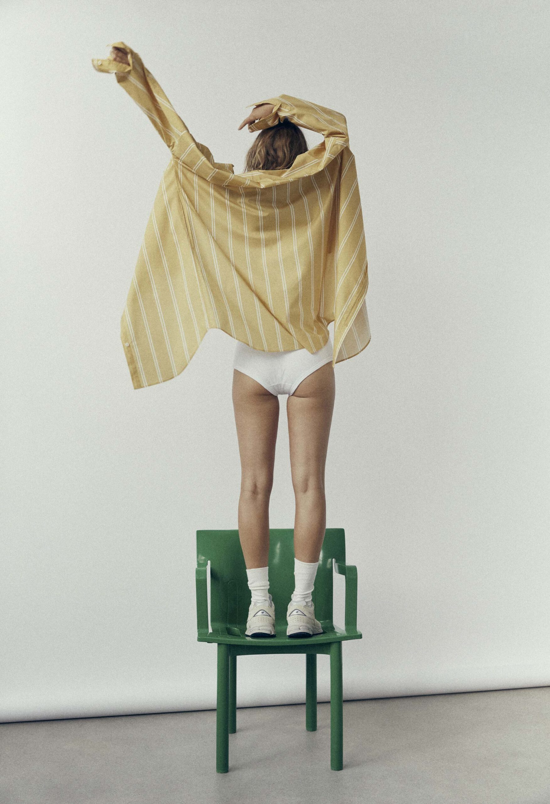 Model standing on a green chair wearing a striped yellow shirt and white shorts against a white backdrop