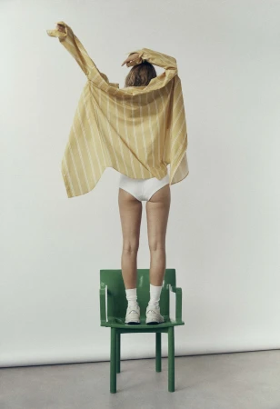 Model standing on a green chair wearing a striped yellow shirt and white shorts against a white backdrop